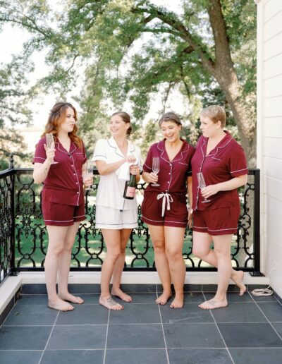 Four women in pajamas stand barefoot on a balcony, holding champagne flutes and a bottle, surrounded by trees in the background.