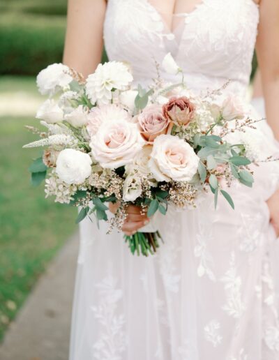 Bride holding a bouquet of white and pink flowers with greenery, wearing a white lace dress in an outdoor setting.