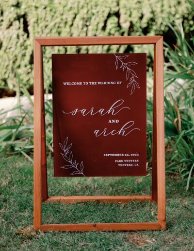 Wedding welcome sign for Sarah and Arch, featuring floral designs, held in a wooden frame on grass.