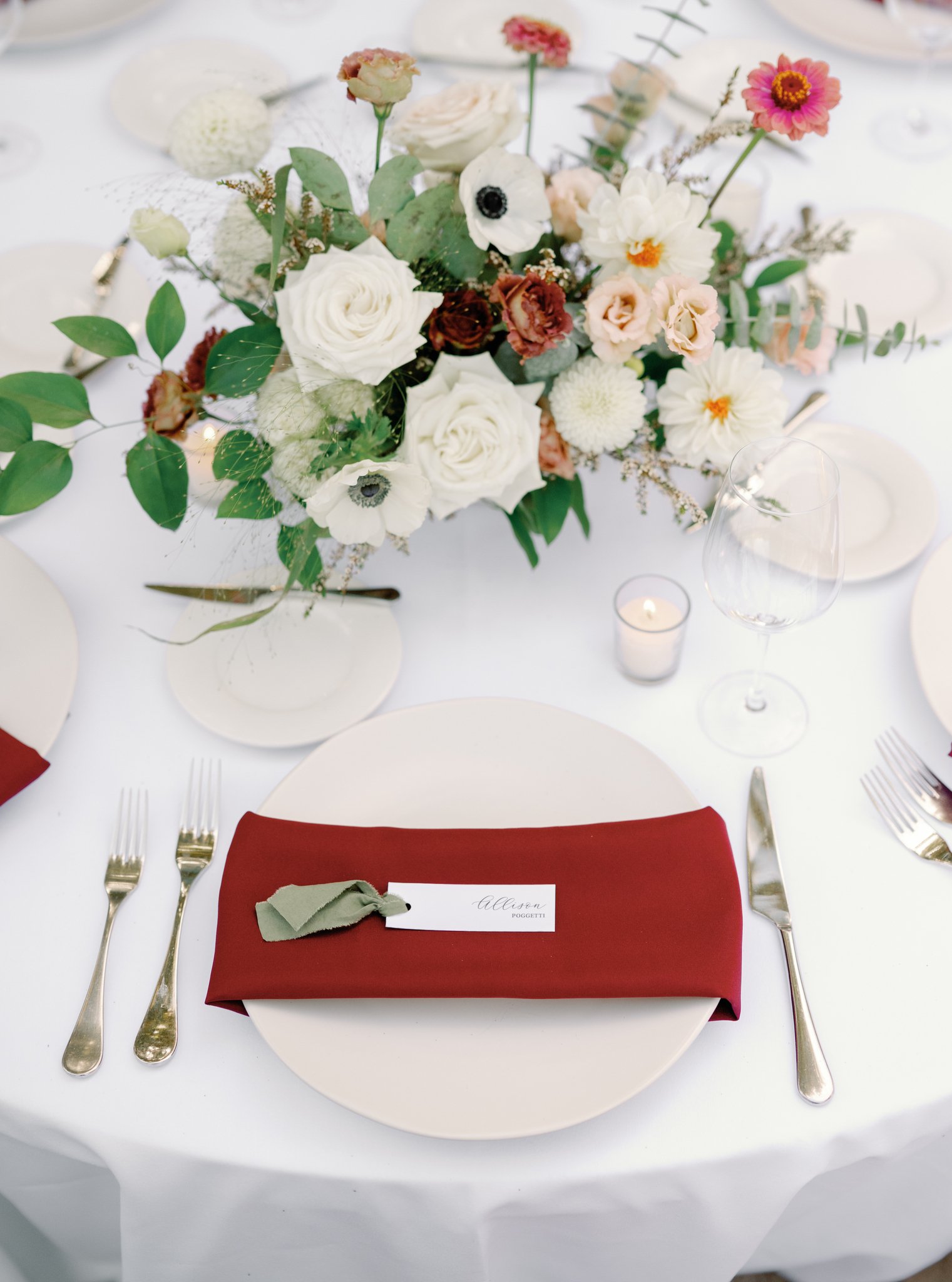 Elegant table setting with a floral centerpiece, gold cutlery, wine glass, and a red napkin with a place card on a white plate.