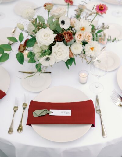 A round table set with a floral centerpiece, white plates, gold cutlery, a wine glass, and a red napkin with a name card.