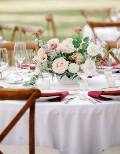 A round table is set for an event with a white tablecloth, floral centerpiece, wine glasses, and folded red napkins on plates. Wooden chairs surround the table.