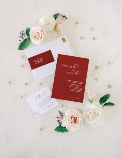 A wedding invitation suite with red and white cards, featuring elegant script and floral accents, surrounded by white and blush roses.