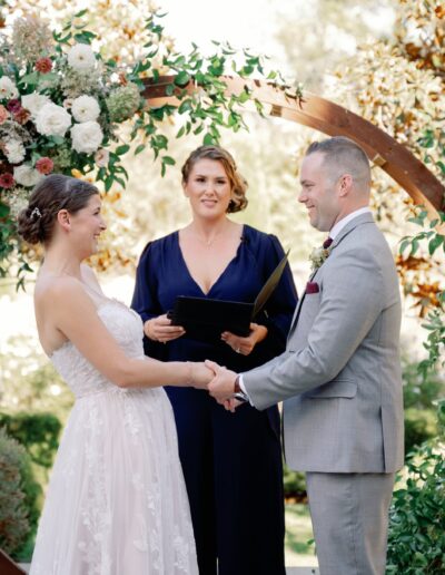 A couple holds hands during their wedding ceremony in front of a floral arch, with an officiant standing between them.