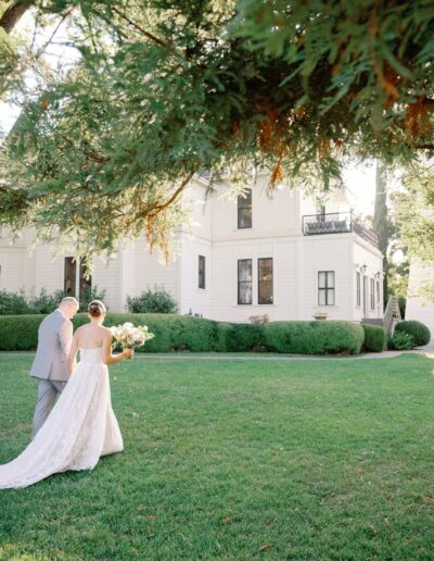 A bride and groom walk across a grassy lawn toward a large white house, surrounded by trees, on a sunny day.