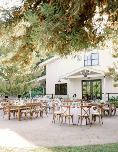 Outdoor wedding reception setup with wooden tables and chairs on a patio, surrounded by trees, near a white barn-like building and a tower.