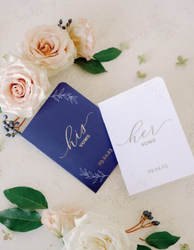 Two vow booklets, one navy labeled "his" and one white labeled "her," are surrounded by pink and white roses and greenery, with a date of 09.24.23 on both.