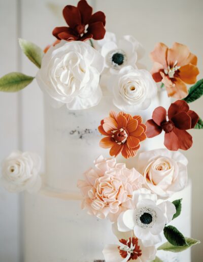 A white tiered cake adorned with realistic-looking edible flowers in shades of white, pink, and red, with green leaves.