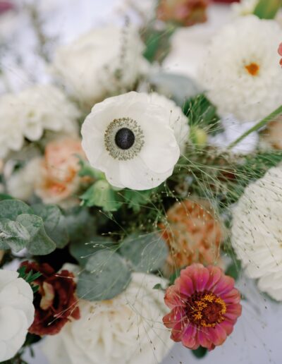 Close-up of a floral arrangement featuring white anemones, pink and red zinnias, and green foliage on a light background.