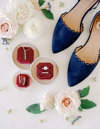 Flat lay of rose flowers, navy blue scalloped shoes, and three open red velvet jewelry boxes displaying a necklace, earrings, and a ring set on a light surface.