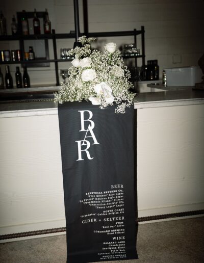 A black bar menu sign with white text stands in front of a bar counter, adorned with white flowers and greenery at the top.