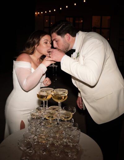 A couple in formal attire drinks from glasses at the top of a champagne tower.