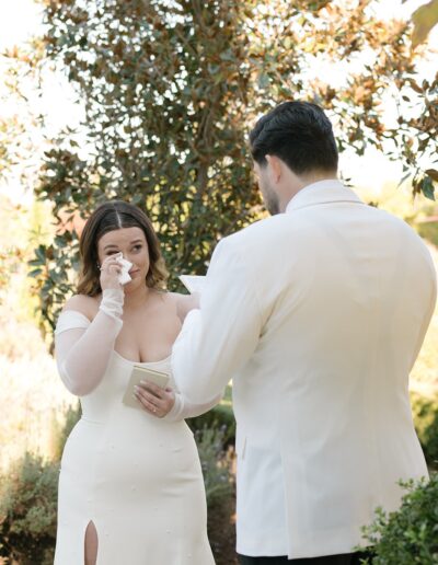 A bride wipes her tears while the groom reads vows during an outdoor ceremony. Both are dressed in white.