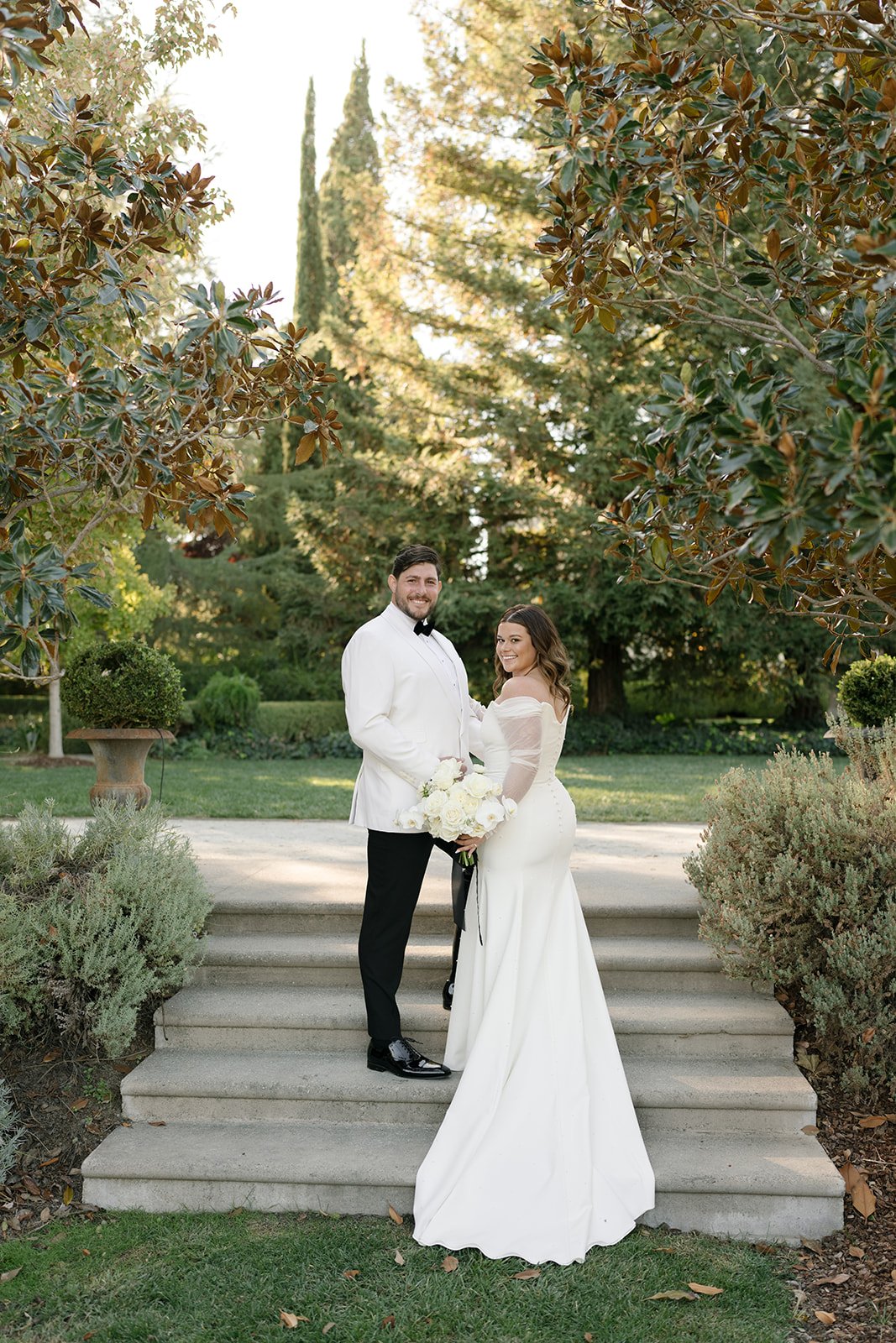 A couple in formal attire stands on outdoor steps surrounded by greenery. The woman holds a bouquet of white flowers, and both look towards the camera.