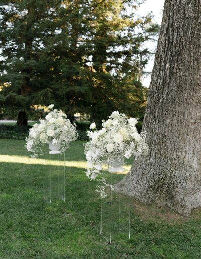 White floral arrangements on stands are placed on grassy ground next to a large tree trunk, with trees in the background.