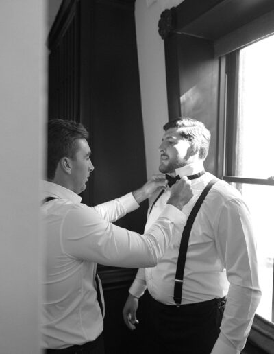 Two men in dress shirts and suspenders, one adjusting the other's bow tie, standing near a window with light streaming in.