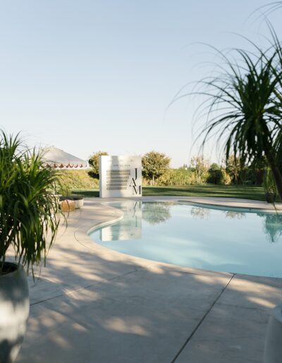 Outdoor swimming pool with surrounding greenery and a white building in the background. Two large potted plants are in the foreground.