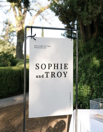 A sign at an outdoor wedding reads "Welcome to the wedding of Sophie and Troy." It is displayed on a metal stand with trees in the background.