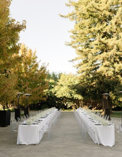 Long outdoor dining setup with two rows of tables covered in white tablecloths and arranged with place settings, surrounded by lush green trees.