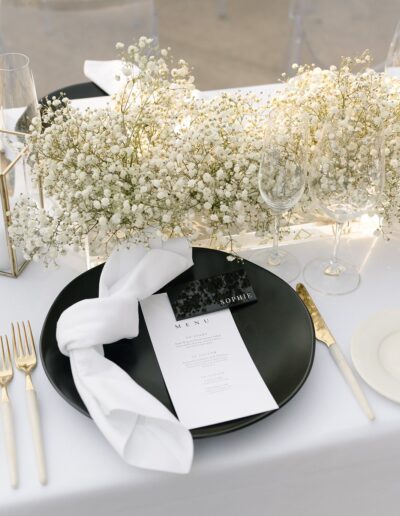 Elegant table setting with a black plate, white napkin, menu, and gold utensils. Delicate white flowers and empty wine glasses are arranged on a white tablecloth.