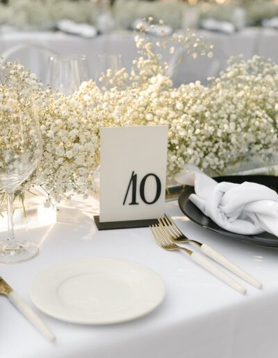 Table setting with a white tablecloth, black plate, white napkin, and gold cutlery. A floral centerpiece with baby's breath surrounds a table number card displaying "10.