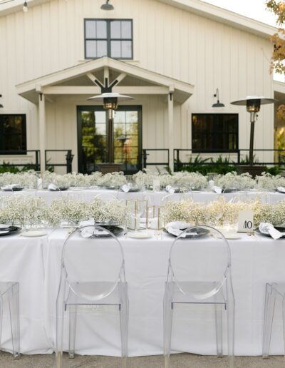 Outdoor event setup with white tablecloths and clear chairs in front of a white building. Tables are decorated with white flowers and candles.