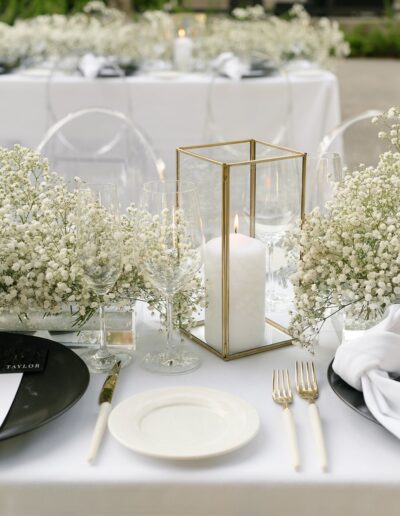 Elegant table setting with white dishes, cutlery, and wine glasses. A lit candle in a glass holder and white flowers decorate the center. Transparent chairs and a white tablecloth are visible.