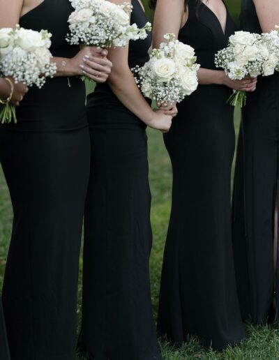 A row of people in black dresses holding white rose and baby's breath bouquets, standing on grass.