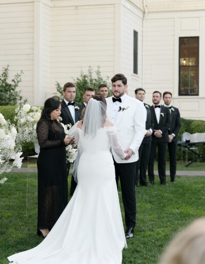 A couple stands in a garden during their wedding ceremony, exchanging vows. The bride wears a white gown and veil, while the groom wears a white tuxedo jacket. Bridesmaids and groomsmen stand nearby.