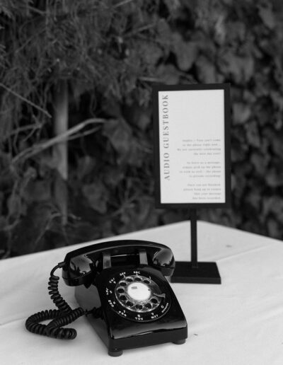 A vintage rotary phone sits on a table next to a sign labeled "Audio Guestbook," set against a backdrop of foliage.