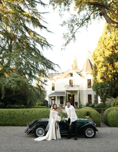 A couple in wedding attire stands beside a vintage black car in front of a large white house surrounded by greenery.