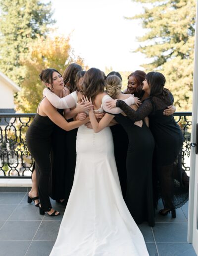 A group of women in black dresses hug a woman in a white dress on a balcony with trees in the background.