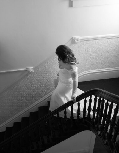 A woman in a white off-shoulder gown descends a staircase, her hand resting on the railing. The setting is indoors with minimal light.