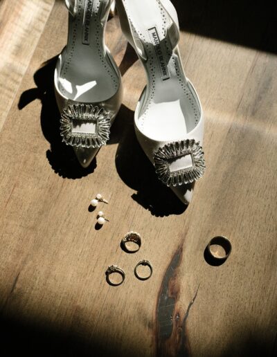 High-heeled shoes with jeweled buckles are placed on a wooden surface alongside pearl earrings and three rings.