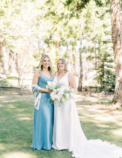 Two women stand on grass in a sunny outdoor setting. One wears a blue dress, the other a white gown holding a floral bouquet. Trees are in the background.