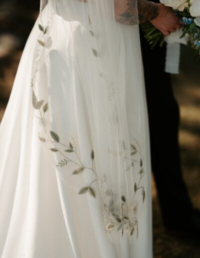 Close-up of a wedding dress with floral embroidery on the veil. The bride holds a bouquet and stands next to another person in black attire.