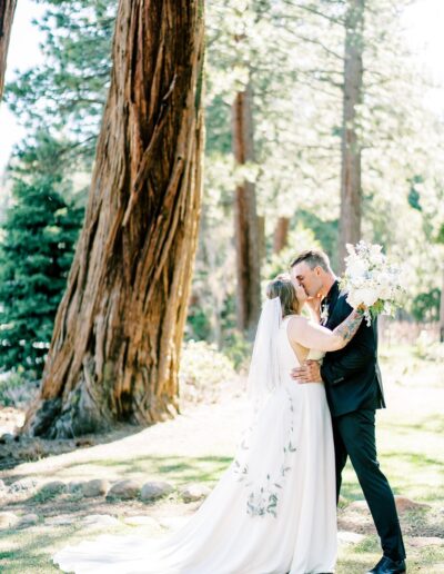 A bride and groom share a kiss outdoors near a large tree, with sunlight filtering through the surrounding woods. The bride holds a bouquet, and both are dressed in formal wedding attire.