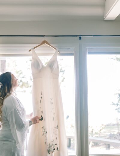 A woman stands in front of a large window, holding a white dress with floral patterns that hangs on a hanger.