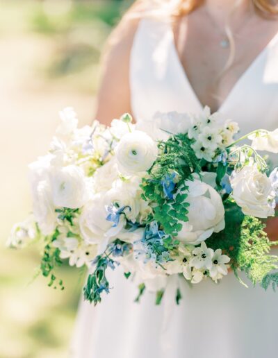 A bride holding a bouquet of white roses, white ranunculus, blue flowers, and ferns against a blurred outdoor background.