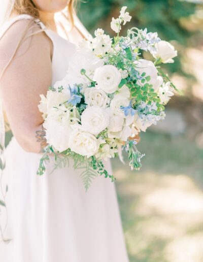 Bride holding a bouquet of white roses, lilies, and blue accents with greenery, wearing a white sleeveless dress in an outdoor setting.