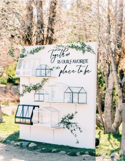 Decorative wall with glass terrariums and greenery, featuring the text "Together is our favorite place to be," outdoors amid leafless trees.