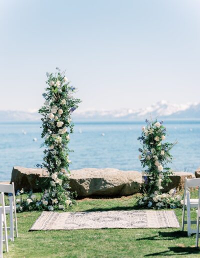 Outdoor wedding altar with floral arrangements by a lake, backed by mountains. Chairs are set up on the grass.