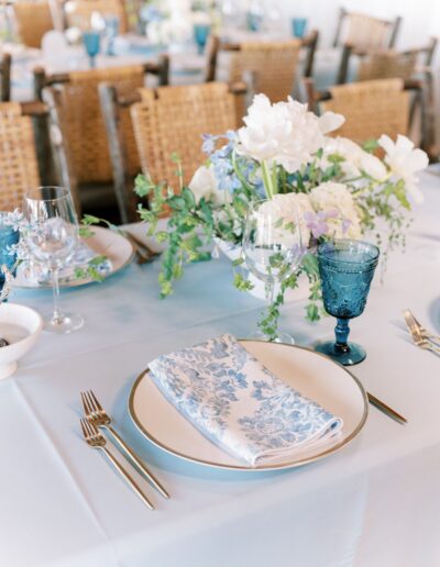 Elegant table setting with a blue and white floral napkin on a plate, surrounded by cutlery, a blue glass, and a floral centerpiece on a light blue tablecloth.