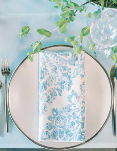 Elegant table setting with a white and blue floral napkin on a plate, surrounded by silverware, a clear wine glass, and a blue water glass, accented by green foliage.