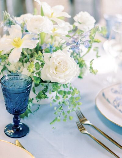 Elegant table setting with white flowers, blue glass goblet, and gold cutlery on a light blue tablecloth.