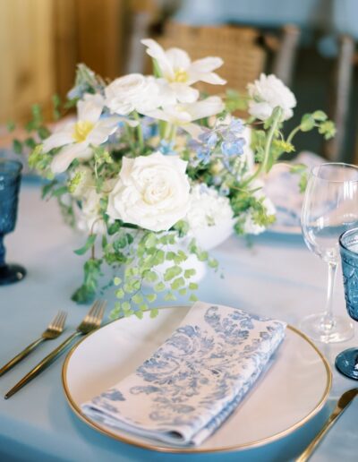 Table setting with a white plate, blue patterned napkin, blue glassware, and a floral centerpiece featuring white and light blue flowers.