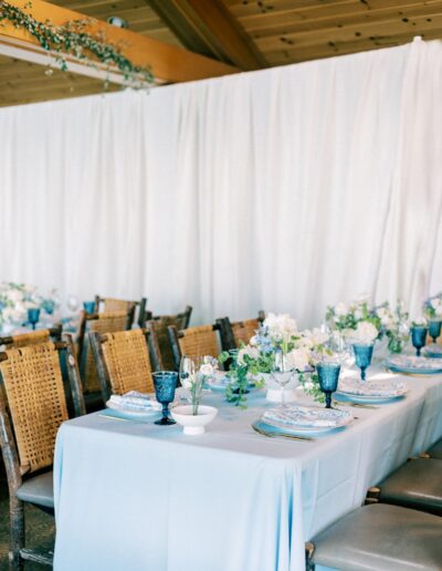 A dining table set with blue tablecloth, woven chairs, glassware, plates, and floral centerpieces, against a white curtain backdrop in a wooden-ceiling venue.