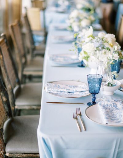 A long dining table with blue tablecloth, set for a meal with blue and white plates, blue glass goblets, floral centerpieces, and wooden chairs.