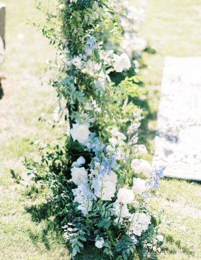 A lush floral arrangement with white roses and light purple flowers, surrounded by green leaves, placed outdoors on grassy ground.