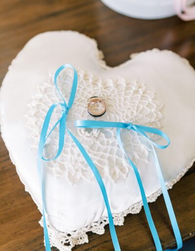 Heart-shaped pillow with lace trim, featuring two blue ribbons and a ring placed on top.